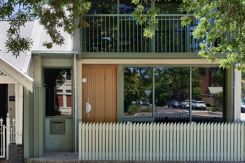 Ground floor entry with vertical corrugated metal fence, timber door, and glazed balcony above among trees