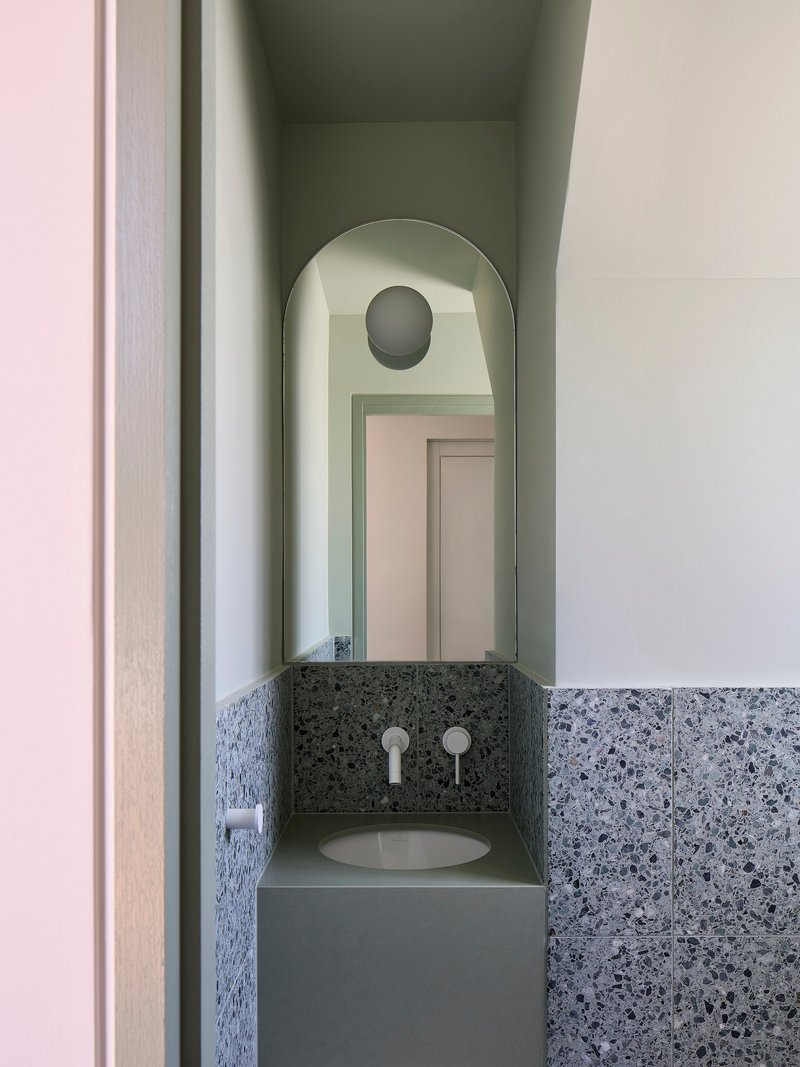 Powder room vanity niche with terrazzo wainscot, round mirror, and arched opening to hallway beyond