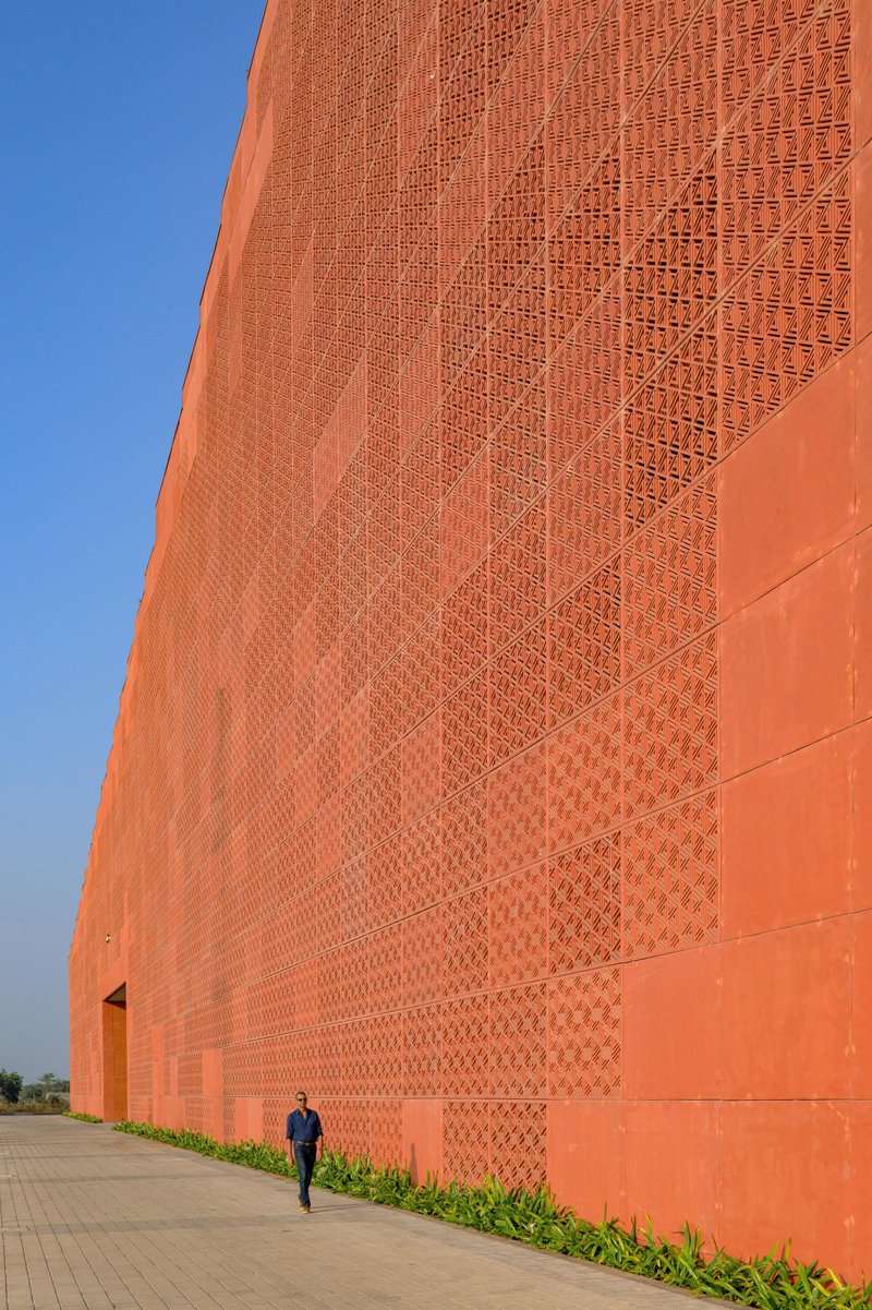 Close-up of orange brick wall with patterned perforations and a figure walking along the paved edge