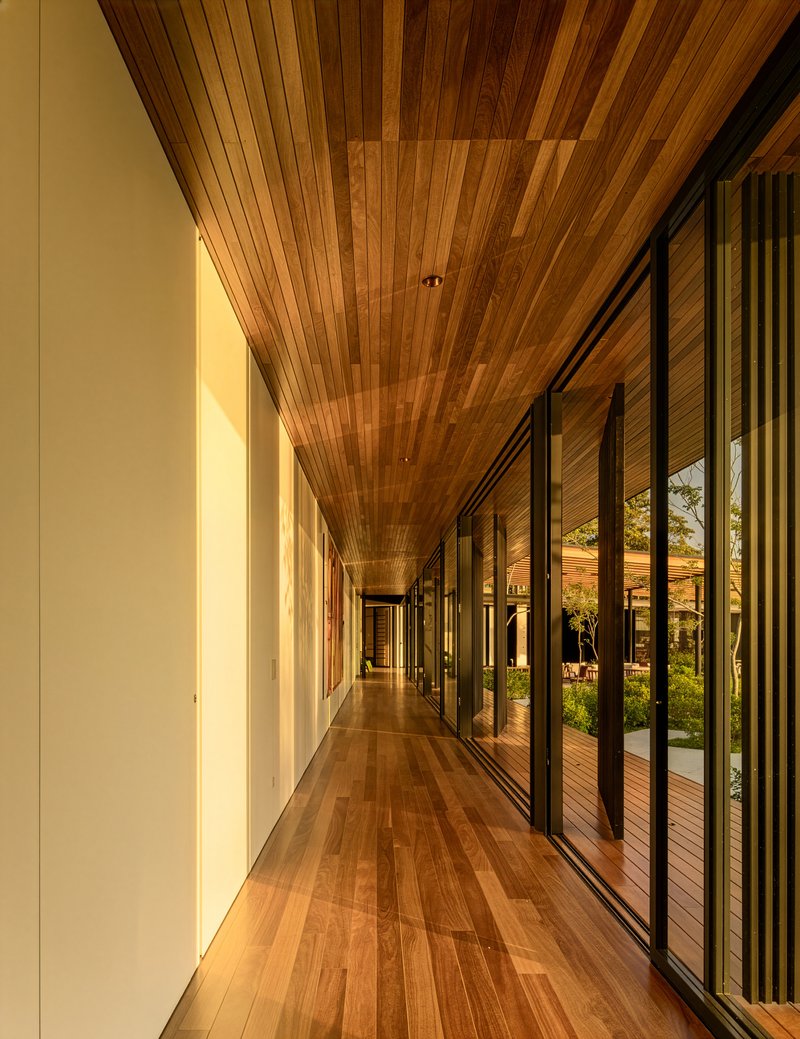Interior corridor with timber plank ceiling and floor-to-ceiling glazing overlooking a courtyard at dusk