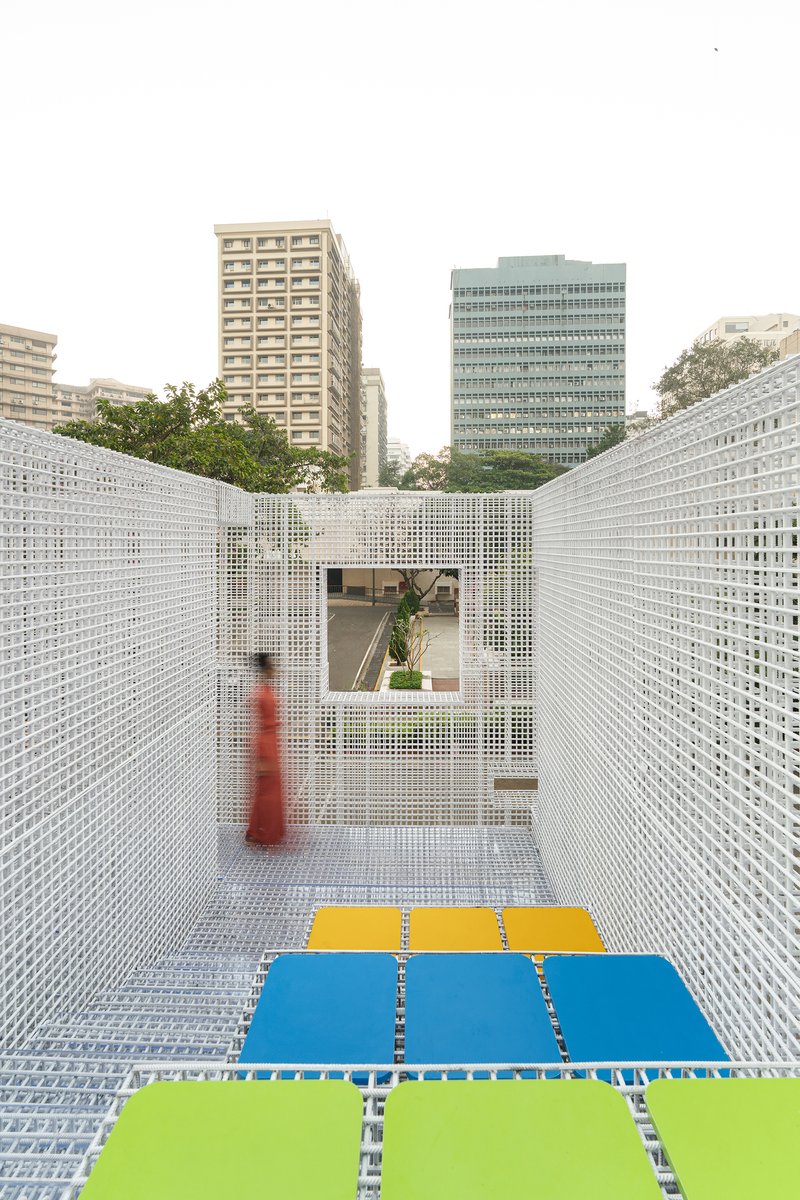 White perforated metal screen corridor with colored floor panels and a visitor in red framed by urban towers