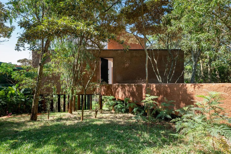 Ground-level view of the brick volume on black pilotis flanked by young trees and dappled sunlight