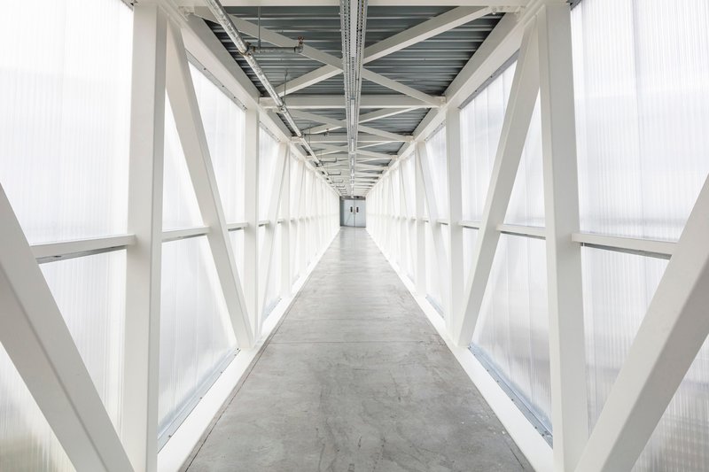 Enclosed corridor with white diagonal steel bracing and translucent polycarbonate cladding under an overcast sky