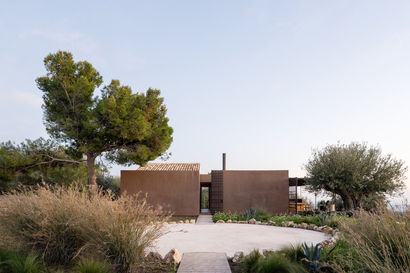 Entrance facade with rammed earth walls framed by ornamental grasses and pine trees