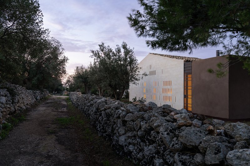 White plastered facade with vertical slit windows glowing at dusk along a dry stone wall path
