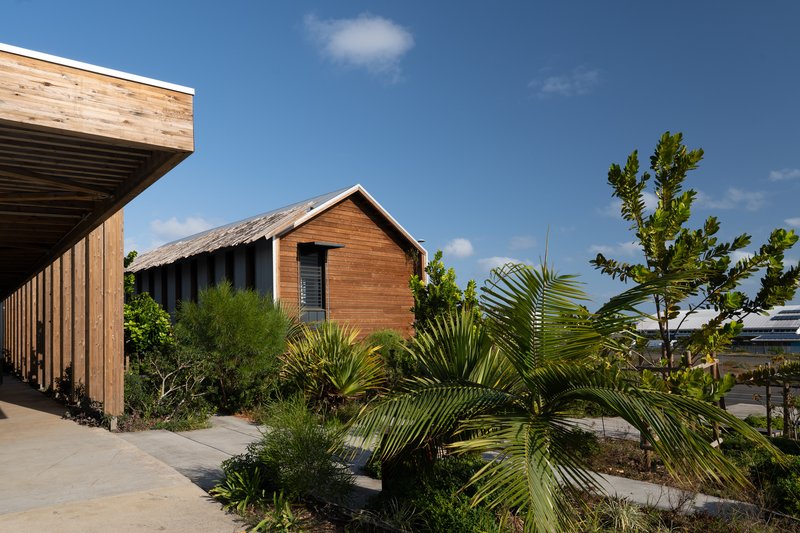Gabled timber building beyond planted bed with palms and tropical vegetation under scattered clouds