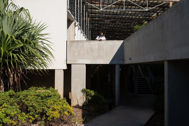 Concrete terrace with overhead trellis and two figures standing on elevated balcony above shadowed passage