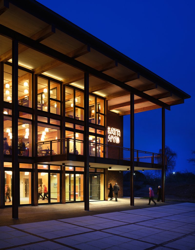 Three-story glass facade with timber ceiling and steel columns lit warmly at blue hour