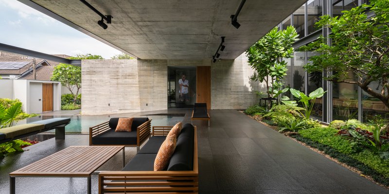 Covered outdoor lounge beneath exposed concrete soffit with timber furniture adjacent to lap pool and planted beds