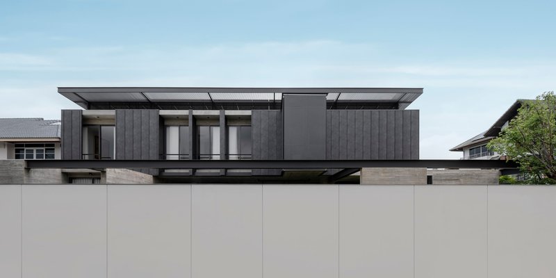 Street view of the dark timber-clad upper volume floating above a beige boundary wall
