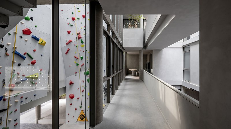 Concrete circulation corridor with colorful climbing wall visible through glass partition and planted courtyard beyond