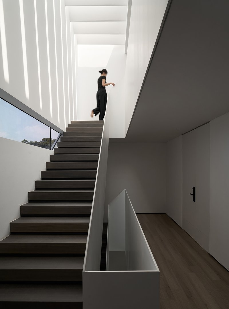 Dark timber staircase with figure ascending under vertical skylights casting striped shadows