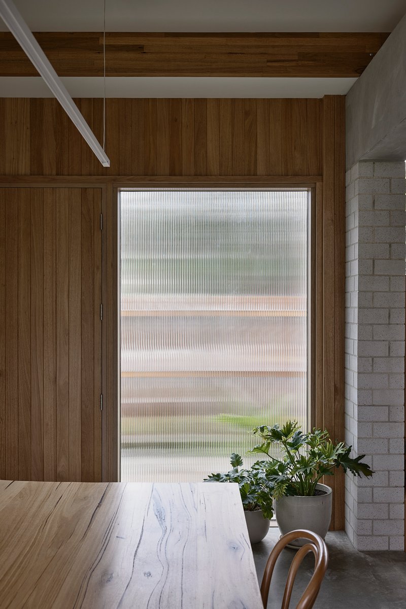 Dining table beneath fluted glass window filtering daylight alongside timber wall and concrete block edge