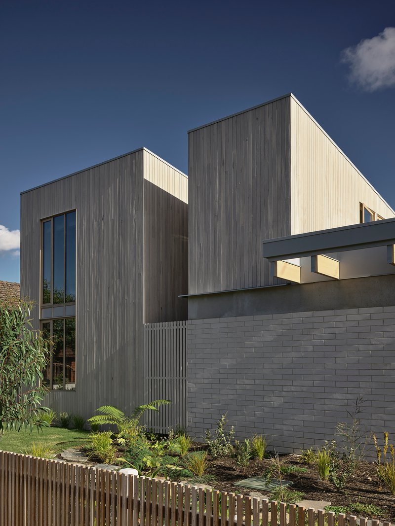 Street facade showing vertical timber cladding above a grey brick base with planted garden bed and timber fence