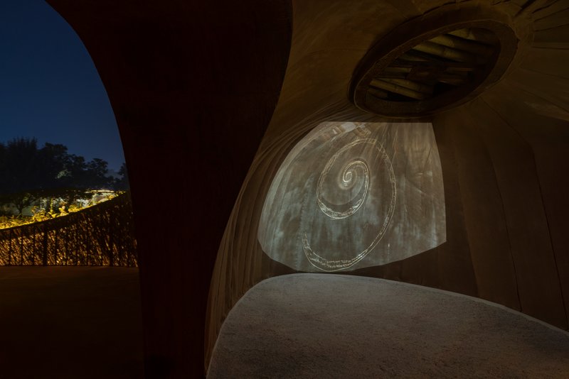 Curved illuminated wall with spiral relief pattern between arched openings and oval roof apertures at dusk