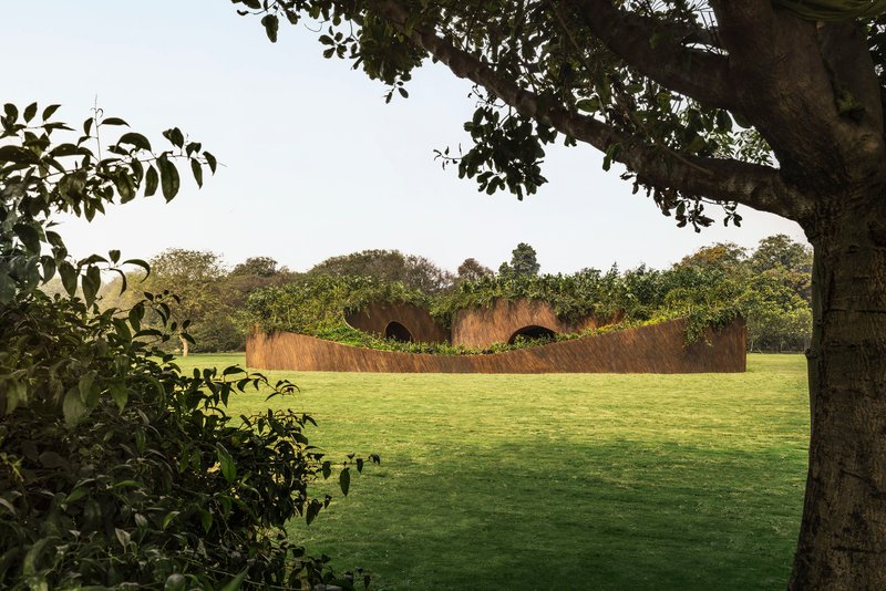 View of the curving weathered steel pavilion across an open lawn framed by tree branches