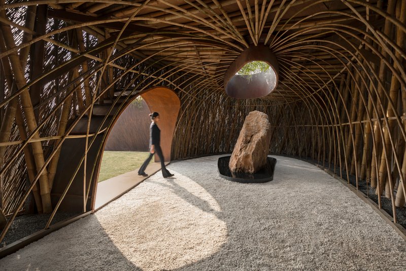 Vaulted bamboo interior structure with figure walking past boulder and gravel floor in dappled sunlight