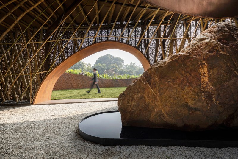Interior view beneath woven bamboo ceiling with large boulder beside reflecting pool as visitor walks through arched opening