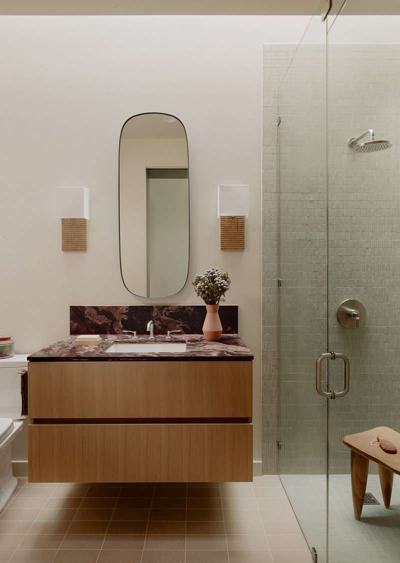 Floating oak vanity with marbled countertop beside a glass-enclosed shower with pale green mosaic tile