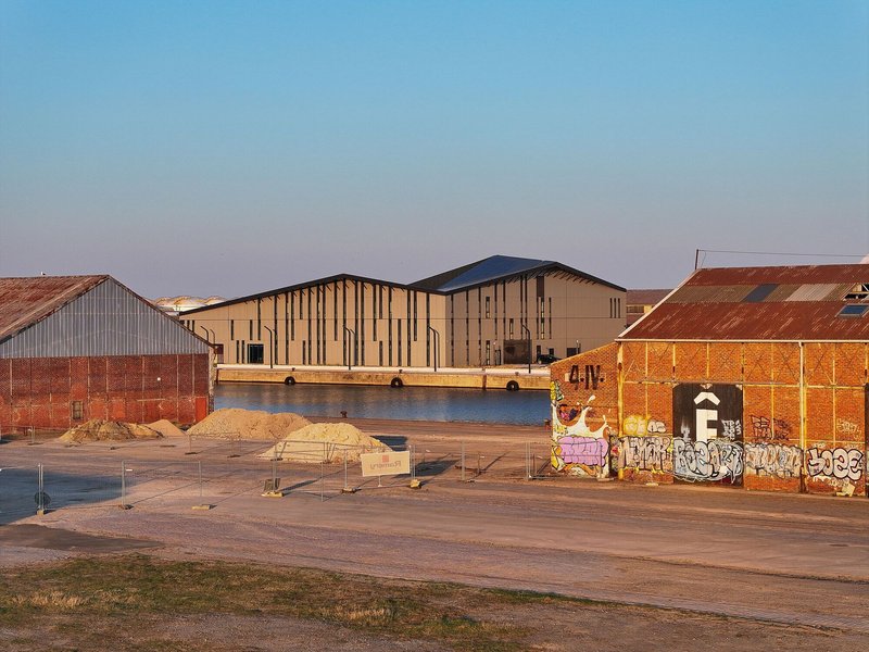 View across the quay showing the zinc roof form rising between graffitied industrial sheds in evening light