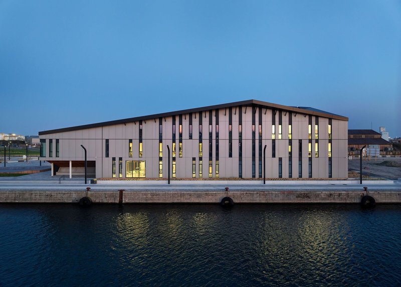 Dockside elevation showing vertical window openings illuminated between timber cladding panels at blue hour
