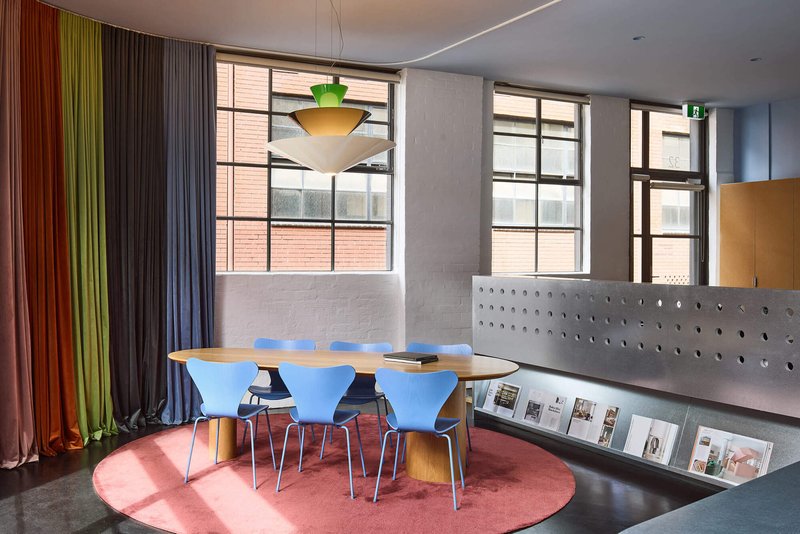 Conference room with oval table and blue chairs on a circular pink rug beneath industrial windows