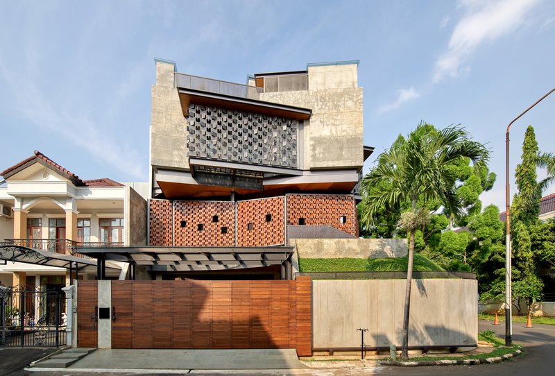 Street view of stacked concrete and perforated brick volumes above a timber fence with palm trees