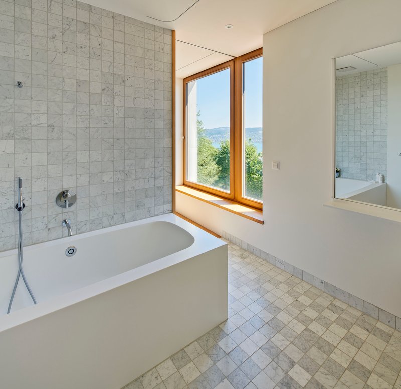 Freestanding tub beneath corner window with pale stone tile wall and distant hills visible outside