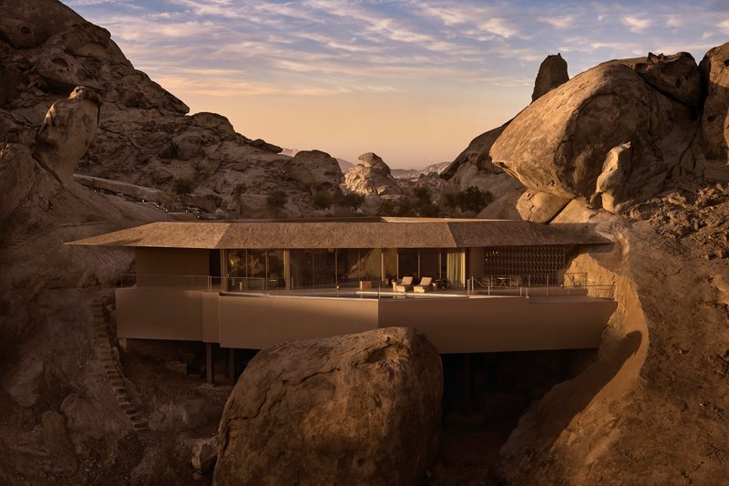Cantilevered concrete terrace with glass walls framed by massive boulders at sunset