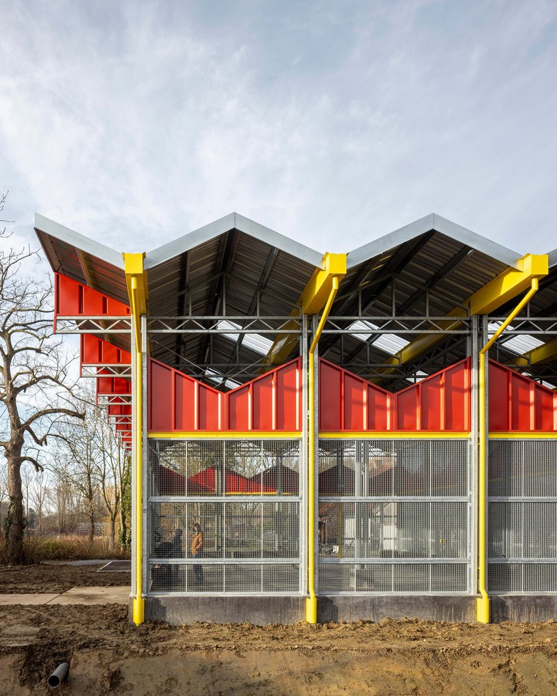 Facade with yellow steel columns, red corrugated panels, and mesh glazing beneath a sawtooth roof on bare ground