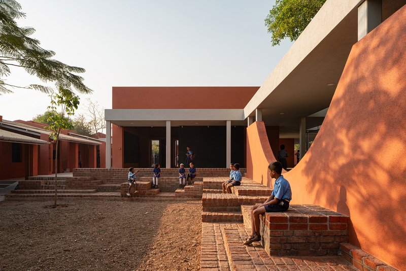 Courtyard with tiered brick seating and children gathering beneath the cantilevered concrete roof in late afternoon sun