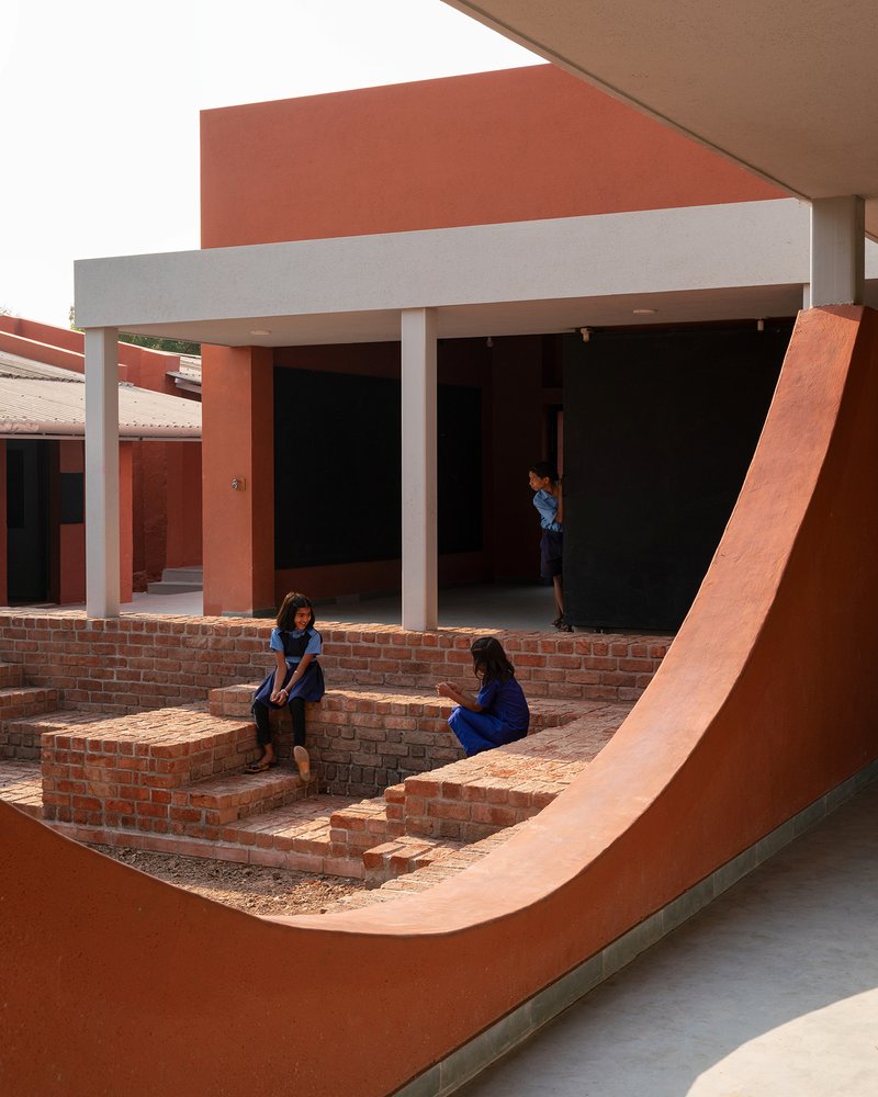 View through the curved terracotta frame toward brick seating steps where children sit in the sheltered courtyard