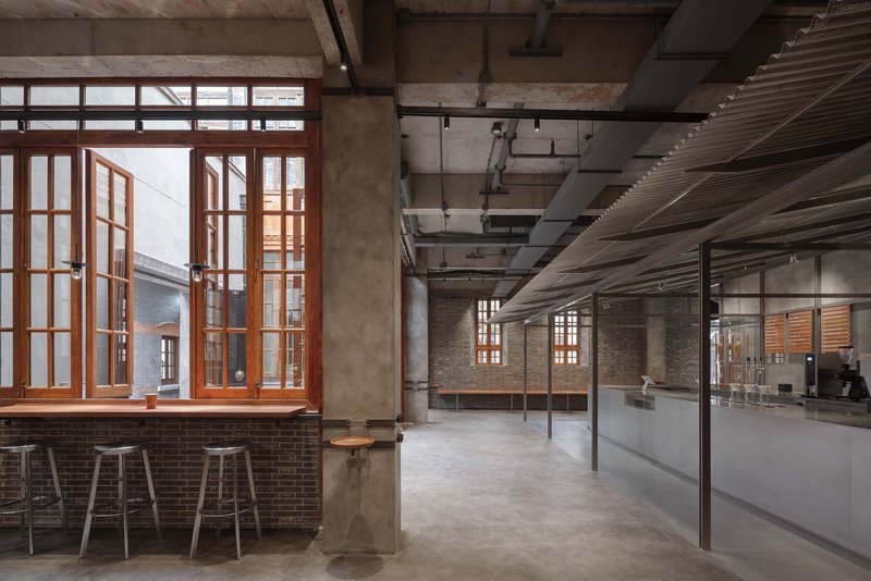 Open interior space with exposed concrete ceiling, timber-framed windows, and brick bar counter with metal stools