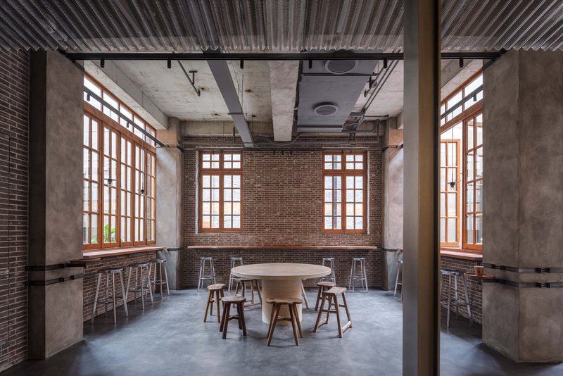 Seating area with timber-framed windows, exposed brick walls, round table and stools beneath corrugated metal ceiling