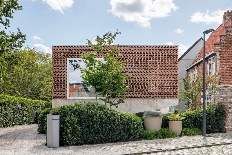 Street view of perforated terracotta brick facade raised on concrete plinth with hedge in foreground