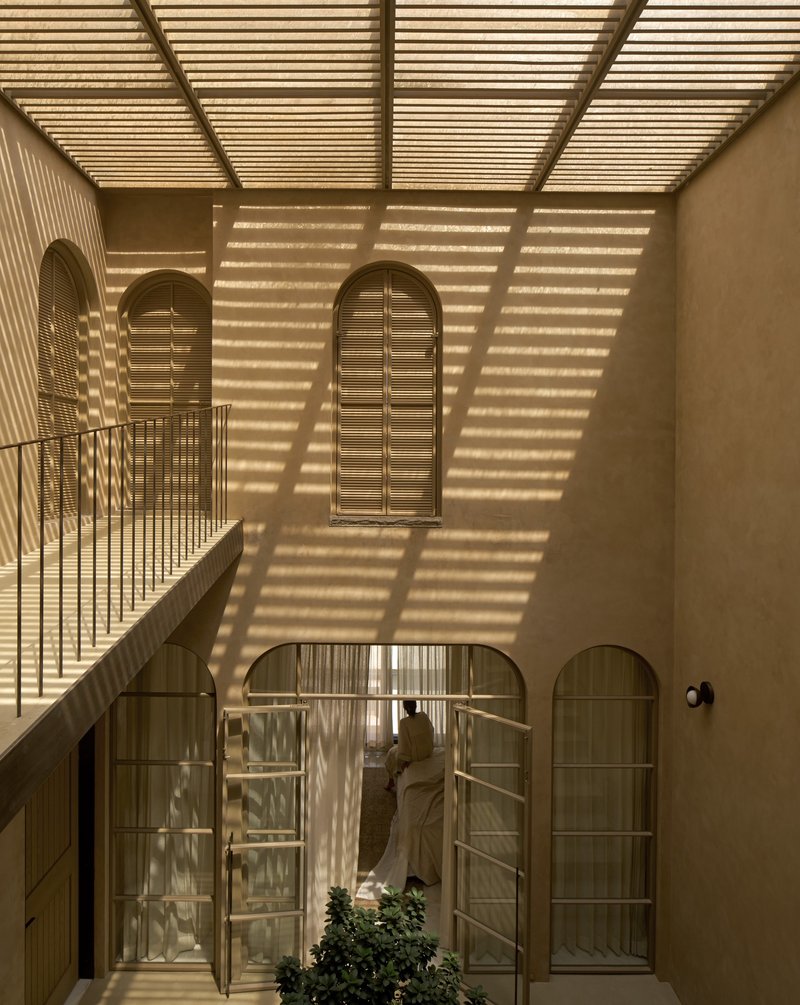 Interior courtyard with arched openings and timber louvered skylight casting striped shadows across plaster walls