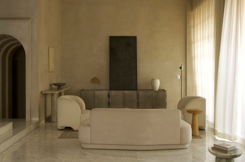 Living room with rounded upholstered furniture, timber credenza, and sheer curtains filtering daylight