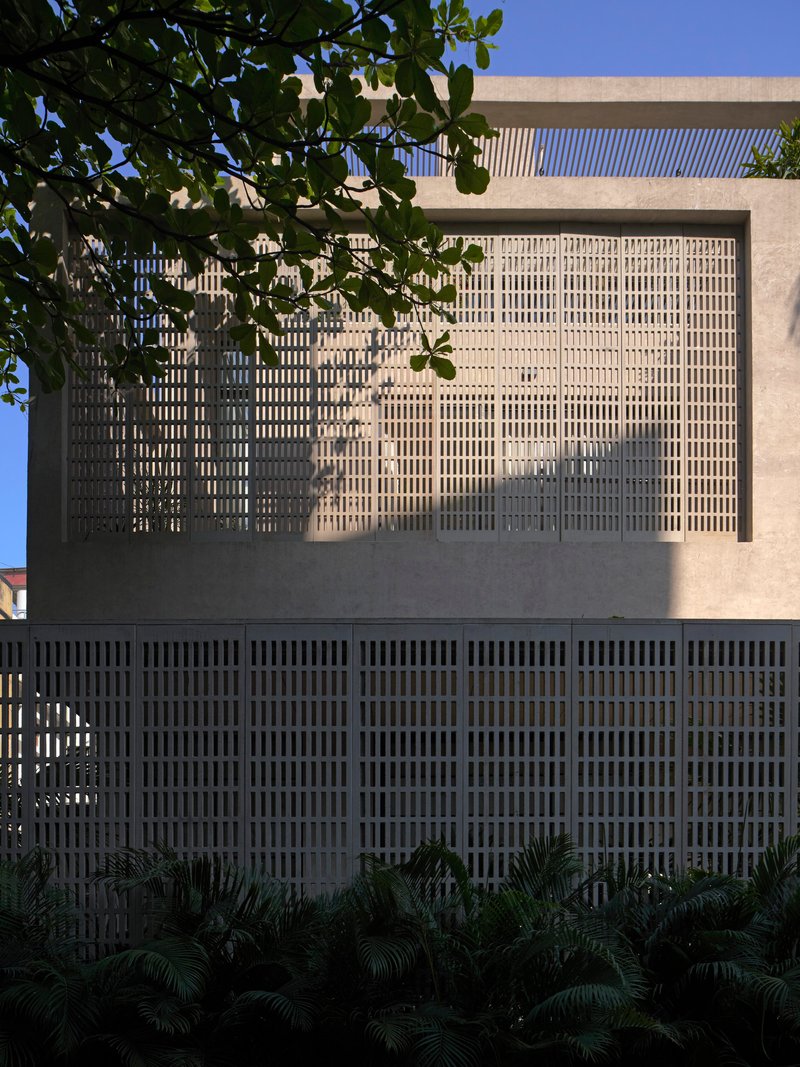 Perforated brick screen facade casting dappled shadows from overhanging foliage onto the concrete frame