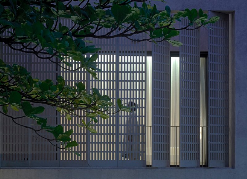 Illuminated vertical-slat facade at dusk with tree branches crossing the perforated concrete screen