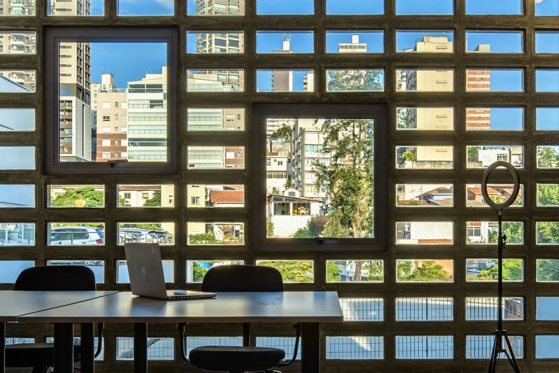 Interior view through concrete grid screen framing a table with laptop overlooking neighboring buildings and trees