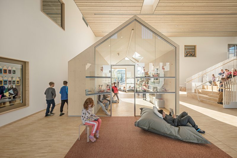 Double-height interior with a glass-enclosed house-shaped pavilion and children on timber stairs