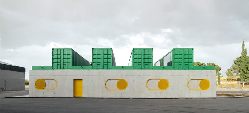 Long concrete facade with paired circular windows and green shipping container modules above under cloudy sky