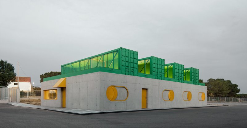 Corner view of concrete volume with circular yellow windows and green shipping containers under grey sky