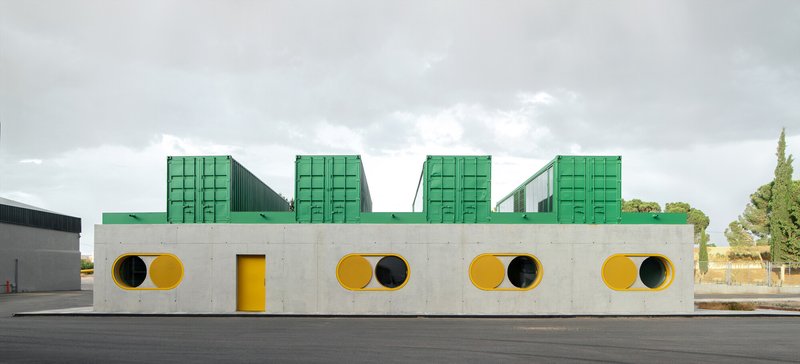 Street-facing concrete facade with paired circular porthole windows and green shipping containers on the roofline