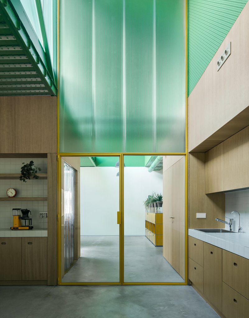 View through yellow-framed glass partition showing kitchen with light wood cabinetry and perforated green ceiling panels