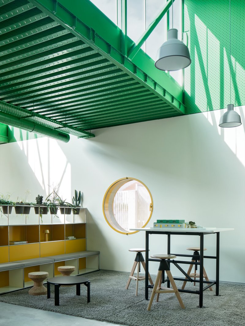 Interior workspace with green perforated metal ceiling structure, round yellow-framed window, and pendant lights