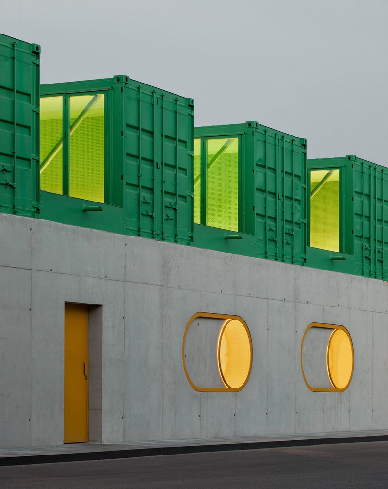 Concrete wall with two circular brass-rimmed windows and yellow door below three green shipping containers with yellow mesh panels