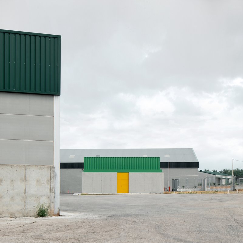 Warehouse facade with green shipping container accent and yellow door in paved yard under cloudy sky