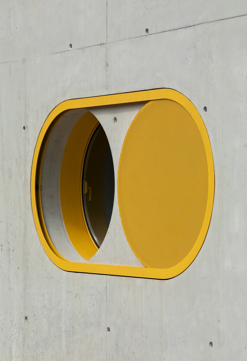 Yellow rounded square window frame with perforated panel set into board-formed concrete wall
