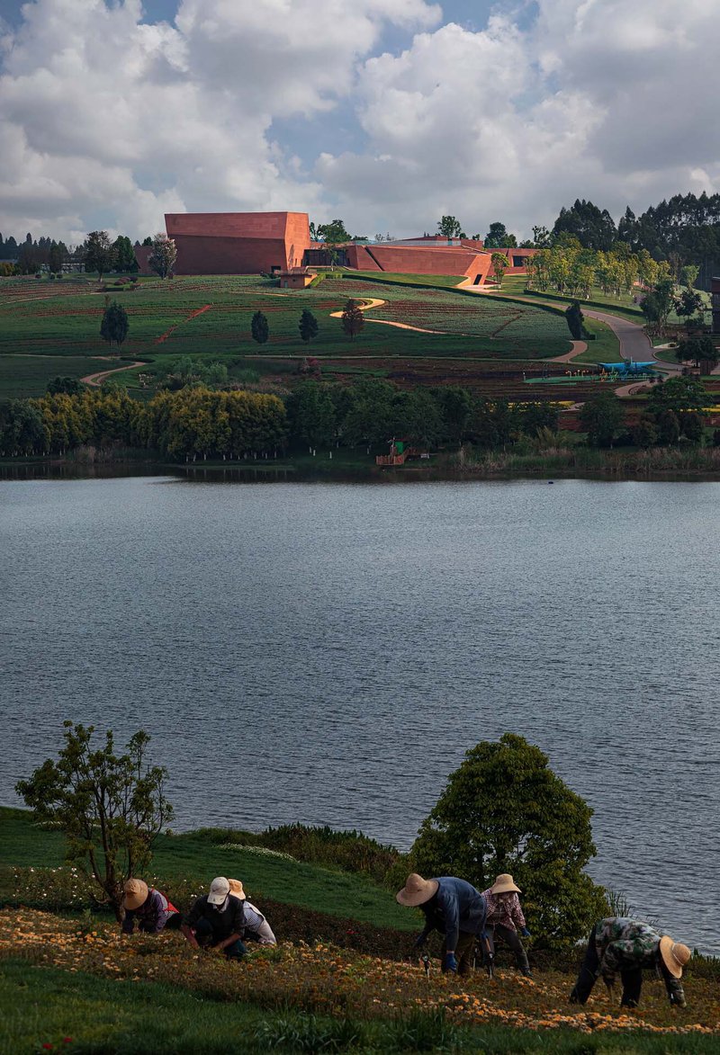 Red earth-toned volumes on terraced hillside with lake in foreground and workers tending the slope